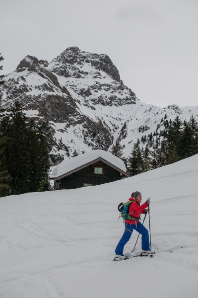 <p>Postkartenmotiv: Skitourengeherin vor schneebedeckter Hütte mit Bergkulisse im Hintergrund. </p>